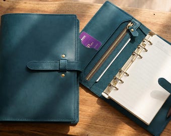 May include: A teal leather planner with a strap closure, featuring a six-ring metal binder mechanism, internal card slots, a zippered pocket, and a pen loop holding a brown pen, with lined white paper pages inserted inside the open organiser.