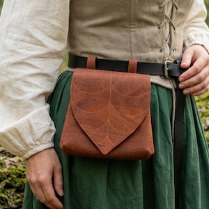 May include: A brown leather belt pouch with a pointed flap and a subtle leaf pattern embossed on the surface. The pouch is attached to a black leather belt worn over a beige lace-up bodice and a long green skirt, creating a functional accessory.