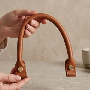 Puede incluir: Un asa de bolso de cuero color canela, curvada y con una textura suave y redondeada. El asa presenta dos puntos de fijación planos y rectangulares en cada extremo, asegurados con un remache metálico circular de tono dorado para un acabado limpio.