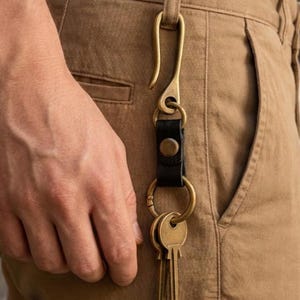 May include: A brass hook keychain attached to a belt loop, featuring a black leather strap with a brass snap button, a circular brass ring, and several brass keys hanging from the bottom, all secured to tan cotton trousers.