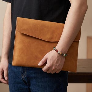 May include: A tan, rectangular document sleeve with a smooth, matte finish and a wide, envelope-style flap closure. The sleeve is held horizontally, showcasing its clean, minimalist design and uniform, warm-toned surface texture.