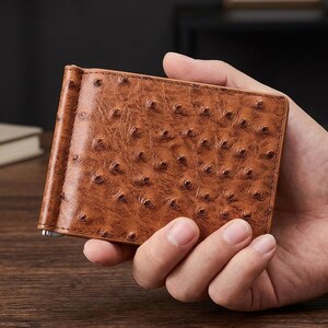 Peut inclure: Un porte-monnaie à pince en cuir marron présentant une texture distincte avec des follicules de plumes en relief. Cet accessoire rectangulaire possède une finition polie et un mécanisme de pince métallique sur le côté gauche pour les billets.