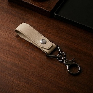 May include: A beige leather keychain strap featuring a silver-toned metal snap button closure. The strap is attached to a dark gunmetal-coloured triangular ring and a matching swivel lobster clasp, providing a secure attachment point for keys.