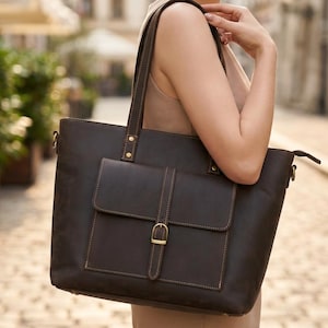 May include: A dark brown leather tote bag featuring two shoulder straps, a rectangular front pocket with a flap closure, and a gold-toned metal buckle. The bag is constructed with visible stitching along the edges and includes metal rivets.