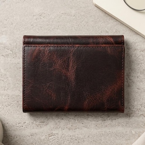 May include: A rectangular, dark brown leather wallet with a visible grain texture and stitched edges. The wallet is folded closed, displaying a smooth, weathered appearance with subtle reddish-brown highlights across the surface of the leather.