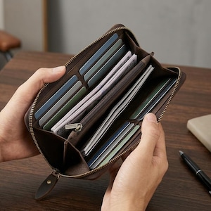 May include: A dark brown leather wallet with a full-length zip closure. The interior features multiple card slots, several open compartments for paper currency, and a central zipped pocket for coins, all organised in a structured layout.