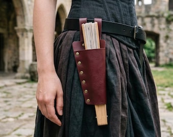 Puede incluir: Una funda para abanico de cuero rojo oscuro con un diseño vertical y remaches metálicos de color latón en el lateral. La funda está sujeta a un cinturón de cuero negro y contiene un abanico de madera plegado con varillas claras.