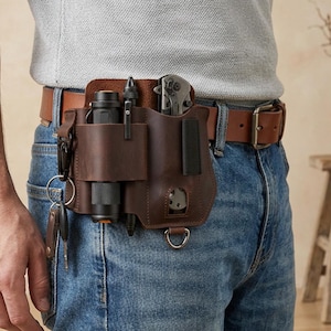 May include: A dark brown leather belt organizer featuring multiple slots for tools, a flashlight, and a folding knife. The organizer is attached to a brown leather belt worn over blue denim jeans and includes a metal D-ring and a key ring attachment.