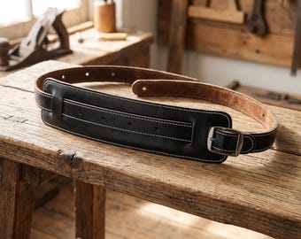 Puede incluir: Una correa de guitarra de cuero negro con una sección acolchada ancha con costuras blancas en contraste, una hebilla de metal plateado y una parte inferior de ante color canela natural. La correa está enrollada y tiene varios agujeros.