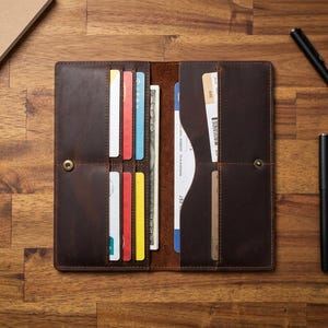 May include: A dark brown leather long wallet featuring multiple vertical card slots, a central compartment for paper currency, and a side pocket for documents. The wallet is secured with a metal snap button closure and contains various colourful cards and bills.