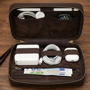 May include: A dark brown leather zipped organiser case with a wrist strap. The interior features multiple pockets and elastic bands holding a white power adapter, a coiled white charging cable, white wireless earbuds, and paper currency.