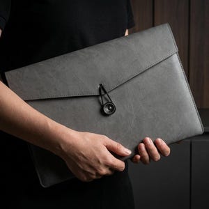 May include: A rectangular gray document folder with a smooth, matte finish and a triangular flap. The folder is secured by a black elastic cord that loops around a circular black button centered on the front flap, featuring visible stitching.
