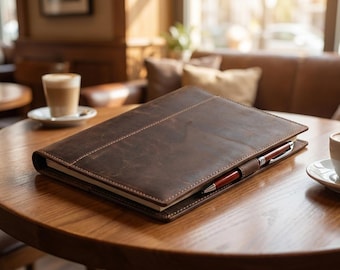 Puede incluir: Una funda de cuero marrón oscuro para cuaderno con costuras visibles en los bordes y una costura central. La funda cuenta con un portalápices que sostiene un bolígrafo fino de color rojo oscuro y plateado, envolviendo el cuaderno.