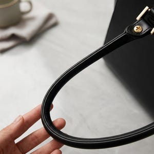 Puede incluir: Un bolso de mano negro con un asa redondeada y lisa sujeta al cuerpo principal mediante herrajes met&aacute;licos dorados. El asa tiene un dise&ntilde;o firme y curvado, y la superficie del bolso presenta un acabado texturizado en color negro.