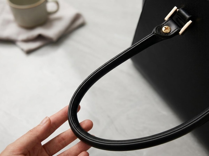 Puede incluir: Un bolso de mano negro con un asa redondeada y lisa sujeta al cuerpo principal mediante herrajes met&aacute;licos dorados. El asa tiene un dise&ntilde;o firme y curvado, y la superficie del bolso presenta un acabado texturizado en color negro.
