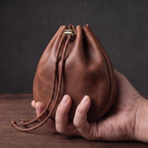 May include: A medium brown leather drawstring pouch with a smooth matte finish. The bag features a rounded base, a cinched top secured by a metallic toggle, and two braided leather cords that hang down from the opening of the pouch.