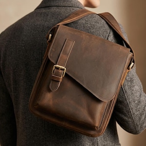Può includere: Una borsa a tracolla in pelle marrone scuro con patta rettangolare, cinturino verticale e chiusura con fibbia in metallo color ottone. La borsa presenta una tracolla larga ed è indossata sopra un blazer grigio testurizzato.