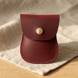 May include: A small, deep red leather pouch with a rounded bottom and a flap closure secured by a single, centered, circular brass-toned metal snap button. The pouch features visible stitching along the edges, creating a structured shape.