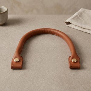 Puede incluir: Un asa curva de color tostado con un agarre redondeado y acolchado. El asa presenta dos puntos de fijaci&oacute;n rectangulares y planos en cada extremo, cada uno asegurado con un remache met&aacute;lico circular y pulido en tono dorado.