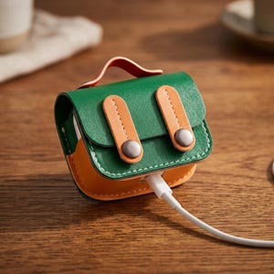 May include: A green and orange leather-like protective case for wireless earbuds, designed to resemble a miniature satchel with a top handle, two vertical straps with silver-toned metal snap buttons, and a white charging cable plugged into the bottom.