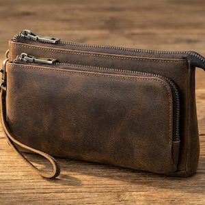Puede incluir: Un bolso de mano de cuero marrón oscuro con dos compartimentos principales con cremallera y un bolsillo frontal adicional con cremallera. El bolso incluye una correa de muñeca extraíble unida a un clip lateral de metal y cremalleras de metal.