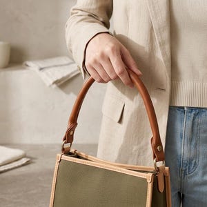 Puede incluir: Un bolso estructurado con cuerpo de lona verde oliva, ribetes de cuero color canela claro y un asa redondeada de cuero marr&oacute;n. El bolso est&aacute; sujeto al asa con herrajes met&aacute;licos dorados y tiene una forma rectangular con costuras visibles.