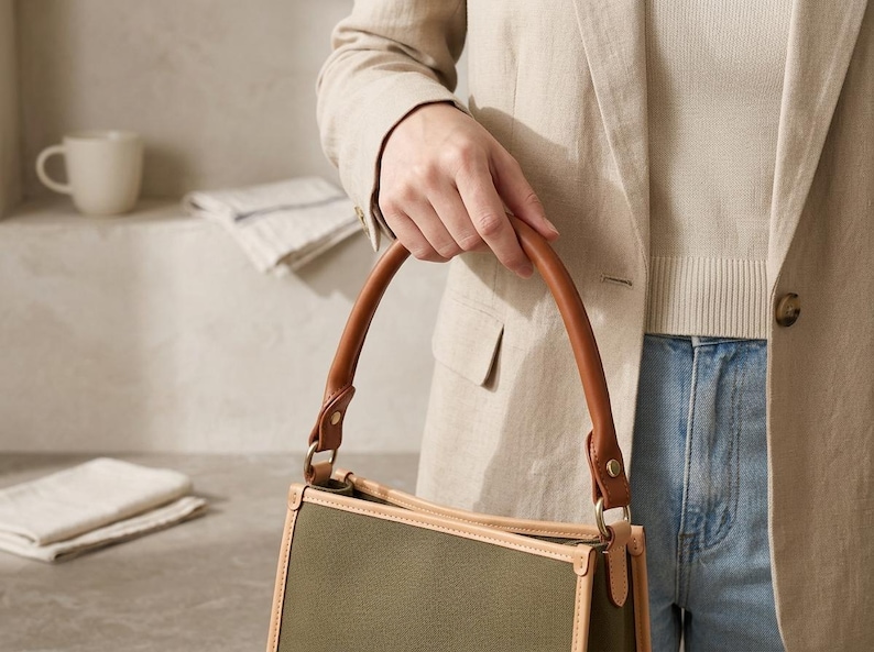 Puede incluir: Un bolso estructurado con cuerpo de lona verde oliva, ribetes de cuero color canela claro y un asa redondeada de cuero marr&oacute;n. El bolso est&aacute; sujeto al asa con herrajes met&aacute;licos dorados y tiene una forma rectangular con costuras visibles.