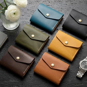 May include: Six small leather wallets in teal, black, olive green, mustard yellow, dark brown, and tan. Each wallet features a triangular flap secured with a single brass-colored metal snap button. The wallets are arranged in a group.