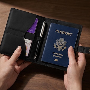 May include: A black leather passport holder featuring a snap button closure, a central pen loop with a silver and black pen, and interior slots containing a blue United States of America passport and two rectangular credit cards with visible text.