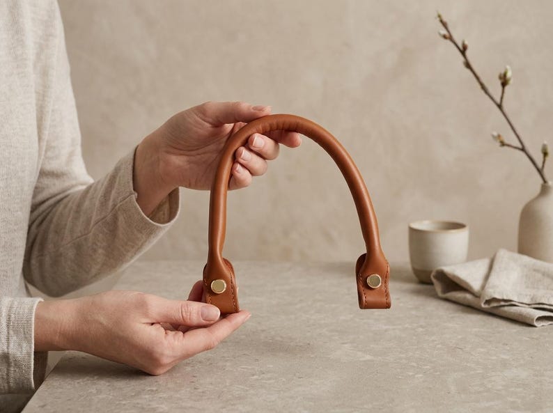 Puede incluir: Un asa de bolso de cuero color canela, curvada y con una textura suave y redondeada. El asa presenta dos puntos de fijaci&oacute;n planos y rectangulares en cada extremo, asegurados con un remache met&aacute;lico circular de tono dorado para un acabado limpio.