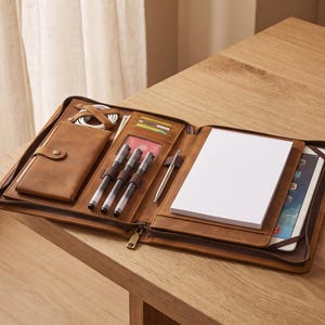 May include: A brown leather zippered portfolio organizer featuring an interior pocket with a snap closure, three pen loops, a card slot, a white notepad, and a tablet sleeve. The organizer is open, displaying its various compartments and storage sections.