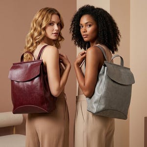 May include: Two backpacks with a fold-over flap closure and a top handle. One backpack is a deep burgundy red, and the other is a muted grey. Both bags feature a smooth, matte finish, a structured base, and adjustable shoulder straps.