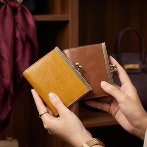 May include: Two rectangular wallets, one in mustard yellow and one in brown, each featuring a metallic frame with a kiss-lock clasp closure. The wallets have a smooth, polished finish and are held together, showcasing their compact design.