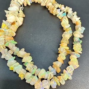 Puede incluir: Un collar de varias hebras compuesto por numerosas pequeñas piezas irregulares de ópalo pulido. Las piedras muestran un aspecto translúcido y lechoso con destellos iridiscentes de color verde, amarillo y naranja en todas las piezas orgánicas.