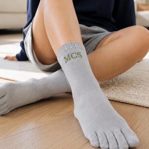 May include: A pair of light gray toe socks featuring individual compartments for each toe. The socks are made of a knit fabric and include the embroidered letters MCS in a dark green thread on the upper portion of the ankle area.