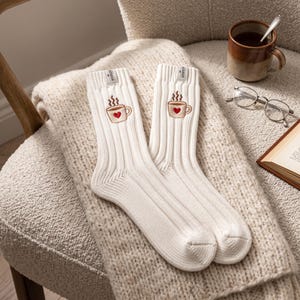 May include: A pair of off-white ribbed crew socks featuring an embroidered graphic of a coffee mug with a small red heart on the front. Each sock has a small rectangular tag at the top cuff with text that appears to read NOBOSSON.