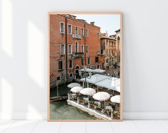 May include: A photograph of a canal in Venice, Italy. The canal is lined with colorful buildings and there are several white umbrellas over tables and chairs at an outdoor cafe. A gondola is in the water.