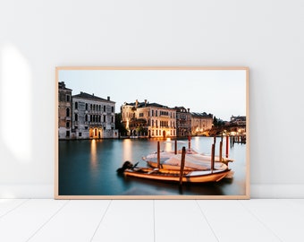 May include: A photograph of a canal in Venice, Italy, with several boats docked along the edge. The buildings on the opposite side of the canal are lit up at dusk, creating a warm glow. The water is calm and reflective, and the sky is a soft blue.