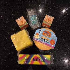 Puede incluir: Una colección de juguetes squishy que incluye un bloque amarillo con agujeros, un recipiente redondo con la etiqueta Squishy Bun Rainbow Mystery, dos cajas pequeñas con personajes, una figura azul brillante y un juguete de mantequilla de 113g.