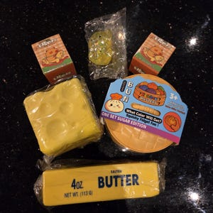 May include: A collection of squishy toys including a yellow block with holes, a round container labeled Squishy Bun Mystery One Set Sugar Edition, a yellow stick labeled 4 oz. Salted Butter, two small boxes with cartoon characters, and a clear plastic bag.