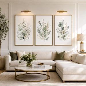 May include: A set of three vertical wall art prints featuring watercolour botanical illustrations of green eucalyptus leaves and dried grasses on a white background, each framed in a thin gold metal frame and illuminated by wall sconces.