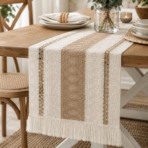 May include: A rectangular table runner featuring a woven texture with alternating vertical stripes in cream and tan. The design includes intricate openwork lace detailing between the stripes and a long, straight fringe along the bottom edge.
