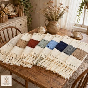 May include: A collection of rectangular crochet table runners featuring a cream-colored base with a central textured band in various colors including tan, rust, sage green, light blue, navy blue, and gray, finished with cream-colored fringe.