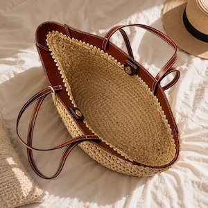 May include: A large open-top tote bag featuring a woven straw body in a natural beige tone, accented with brown leather trim along the upper edges and handles. The bag includes two thin brown leather straps and gold-toned metal snap closures.