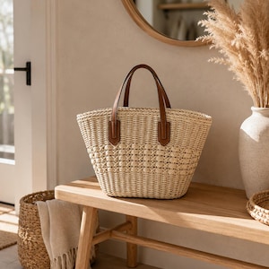 Pode incluir: Uma bolsa tote de palha entran&ccedil;ada em tom bege claro natural, com um padr&atilde;o de cestaria texturizado e duas al&ccedil;as de couro castanho fixadas com abas retangulares. A bolsa possui uma abertura superior e uma silhueta larga e curva.