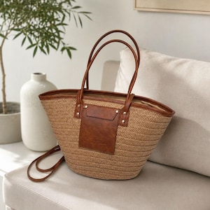 May include: A woven straw tote bag featuring a tan textured body, brown leather-like trim, and a central brown rectangular pocket detail. The bag includes two tall brown handles and a long, thin brown shoulder strap attached to the sides.
