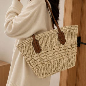 Pode incluir: Uma bolsa tote de palha tran&ccedil;ada bege com um padr&atilde;o entrela&ccedil;ado complexo no centro e uma trama s&oacute;lida na base. A bolsa inclui duas al&ccedil;as de ombro de aspecto couro castanho presas com remendos retangulares na parte externa frontal.