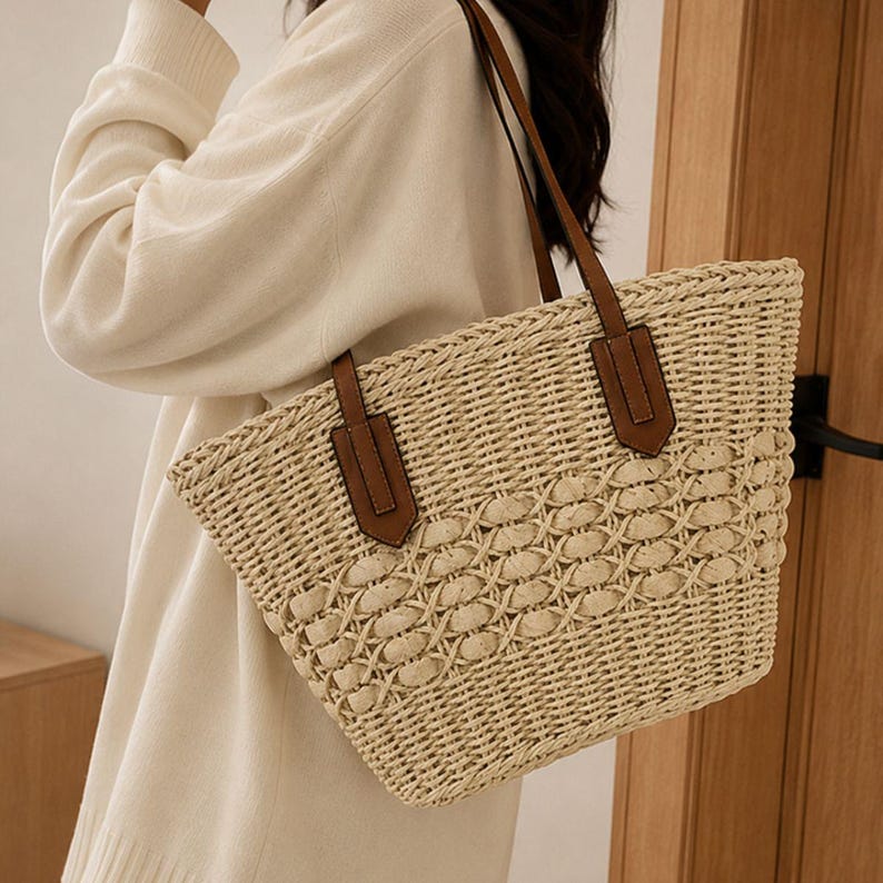 Pode incluir: Uma bolsa tote de palha tran&ccedil;ada bege com um padr&atilde;o entrela&ccedil;ado complexo no centro e uma trama s&oacute;lida na base. A bolsa inclui duas al&ccedil;as de ombro de aspecto couro castanho presas com remendos retangulares na parte externa frontal.