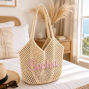 May include: A beige crochet tote bag featuring an open-weave diamond pattern and two long shoulder straps. The front of the bag displays the name Sophia embroidered in raised, light pink cursive lettering across the center of the woven structure.
