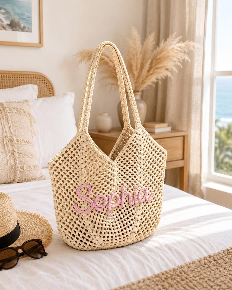 May include: A beige crochet tote bag featuring an open-weave diamond pattern and two long shoulder straps. The front of the bag displays the name Sophia embroidered in raised, light pink cursive lettering across the center of the woven structure.